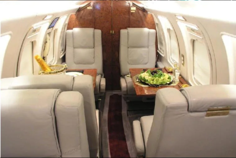 The luxurious interior cabin of a private jet with light grey leather seats, wood tables, and a meal setting.
