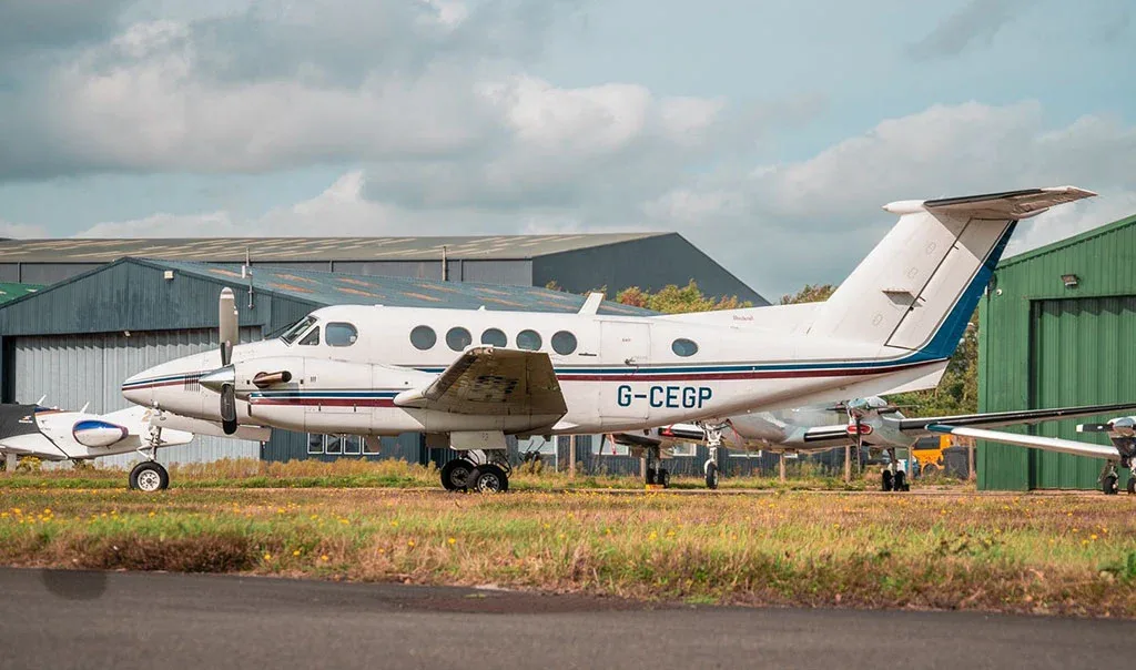 A white twin-turboprop aircraft with blue and red stripes, registration G-CEGP, parked on a grassy area next to hangars.