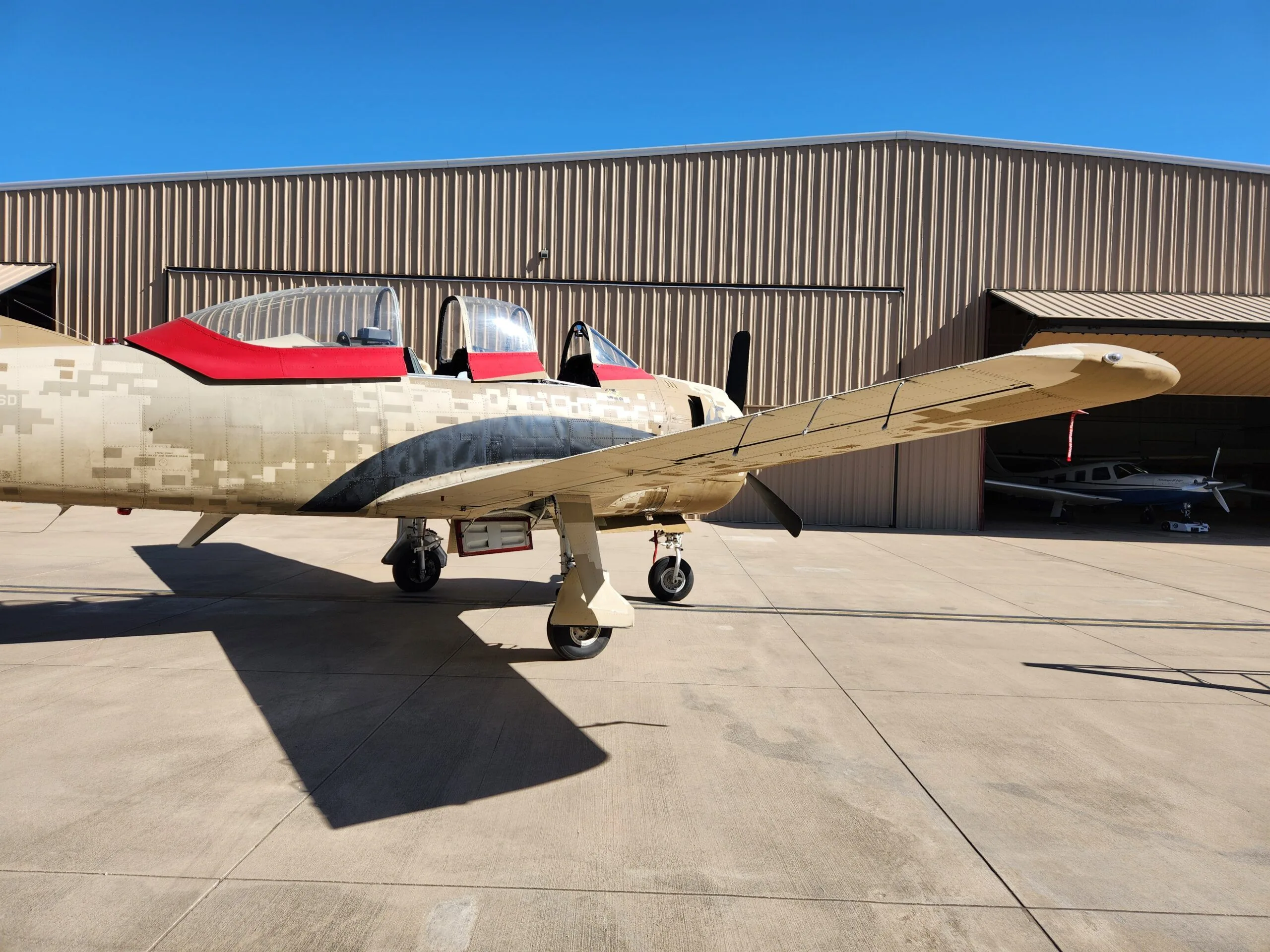 A T-28 Trojan aircraft with desert digital camouflage and red accents is parked on an airfield, with another aircraft visible in a hangar.