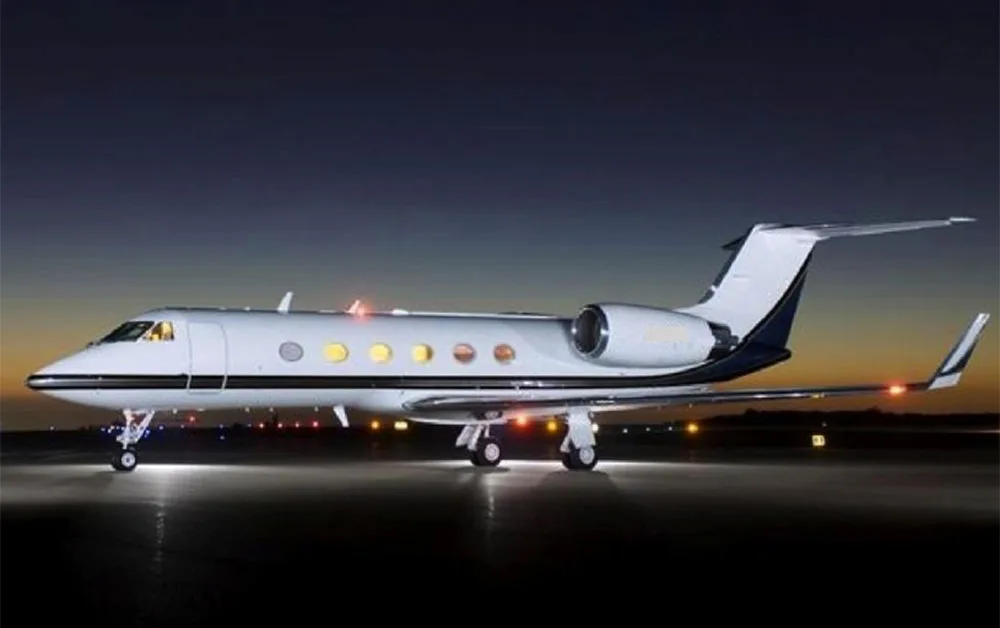 A white Gulfstream G550 business jet with dark blue accents is parked on a tarmac at dusk, illuminated by its own lights and ground lights.