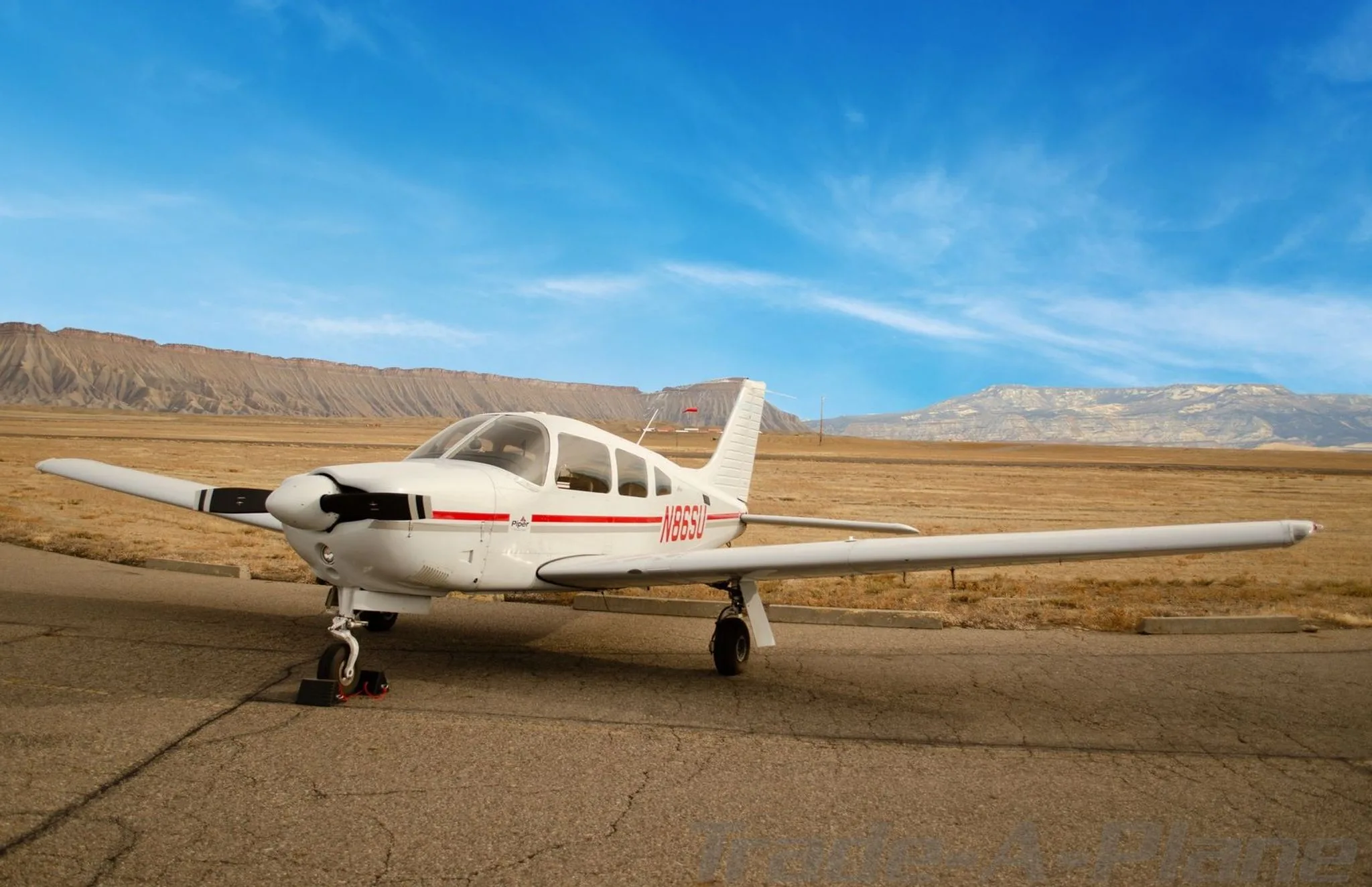 A white Piper aircraft with red stripes and tail number N86SU is parked on an asphalt runway, viewed from the front, with mountains in the background.