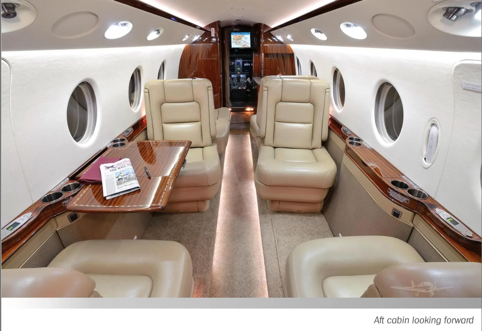A spacious private jet cabin with light-colored leather seats, a wooden table, and a view looking forward towards the cockpit.