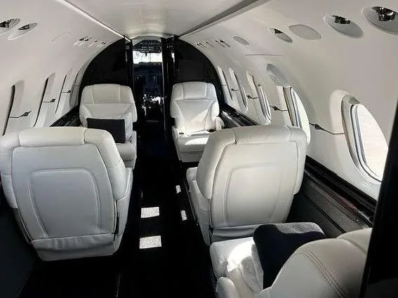 The spacious interior of a private jet cabin, with white leather seats and a view looking towards the cockpit.