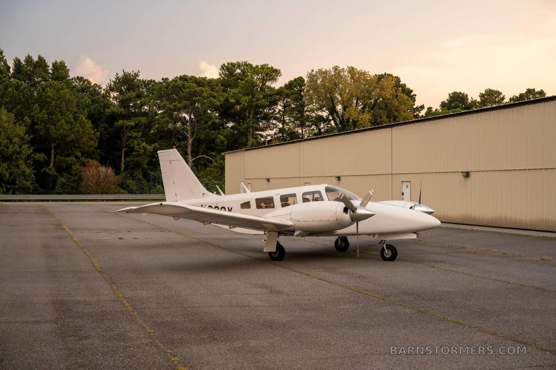 A white twin-engine propeller aircraft (N928X) is parked on an asphalt ramp next to a hangar at sunset.