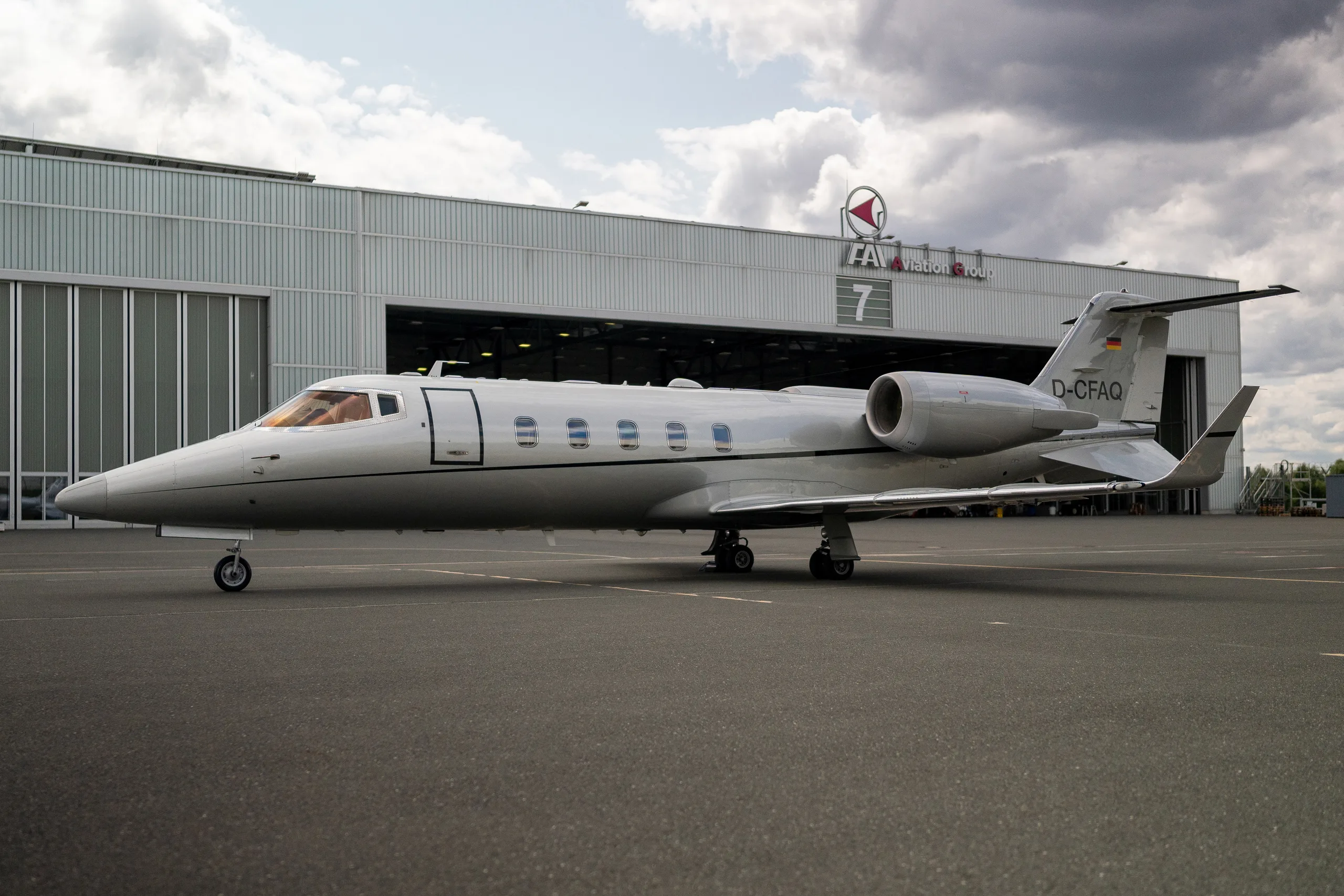 A silver Learjet 60 business jet with registration D-CFAQ parked on a tarmac in front of a hangar under a cloudy sky.