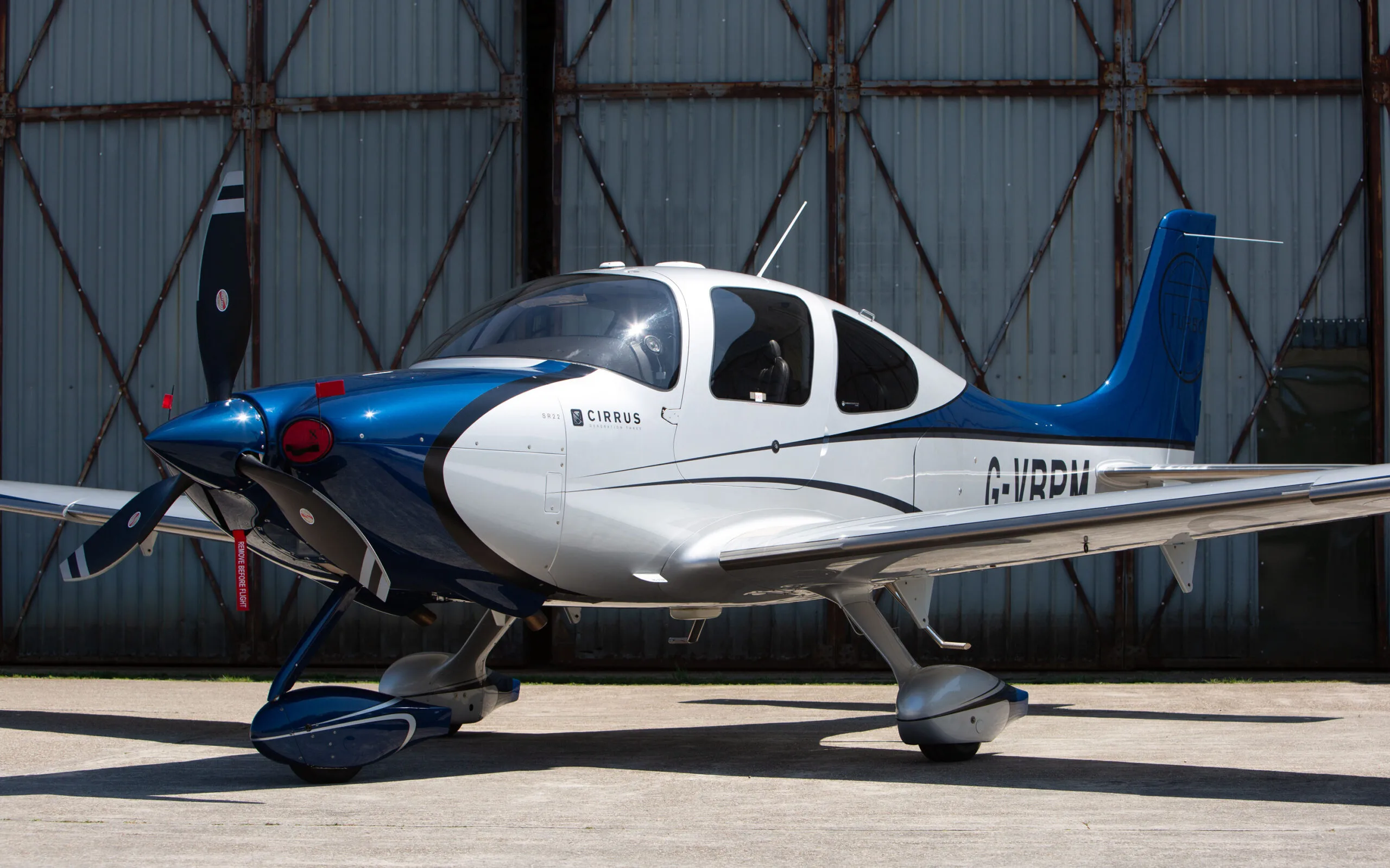 A side view of a blue and white Cirrus SR22 aircraft with registration G-VBPM, parked in front of a hangar.