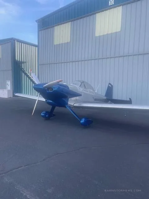 A blue and silver single-engine aircraft with a propeller, parked on an asphalt apron next to hangars.