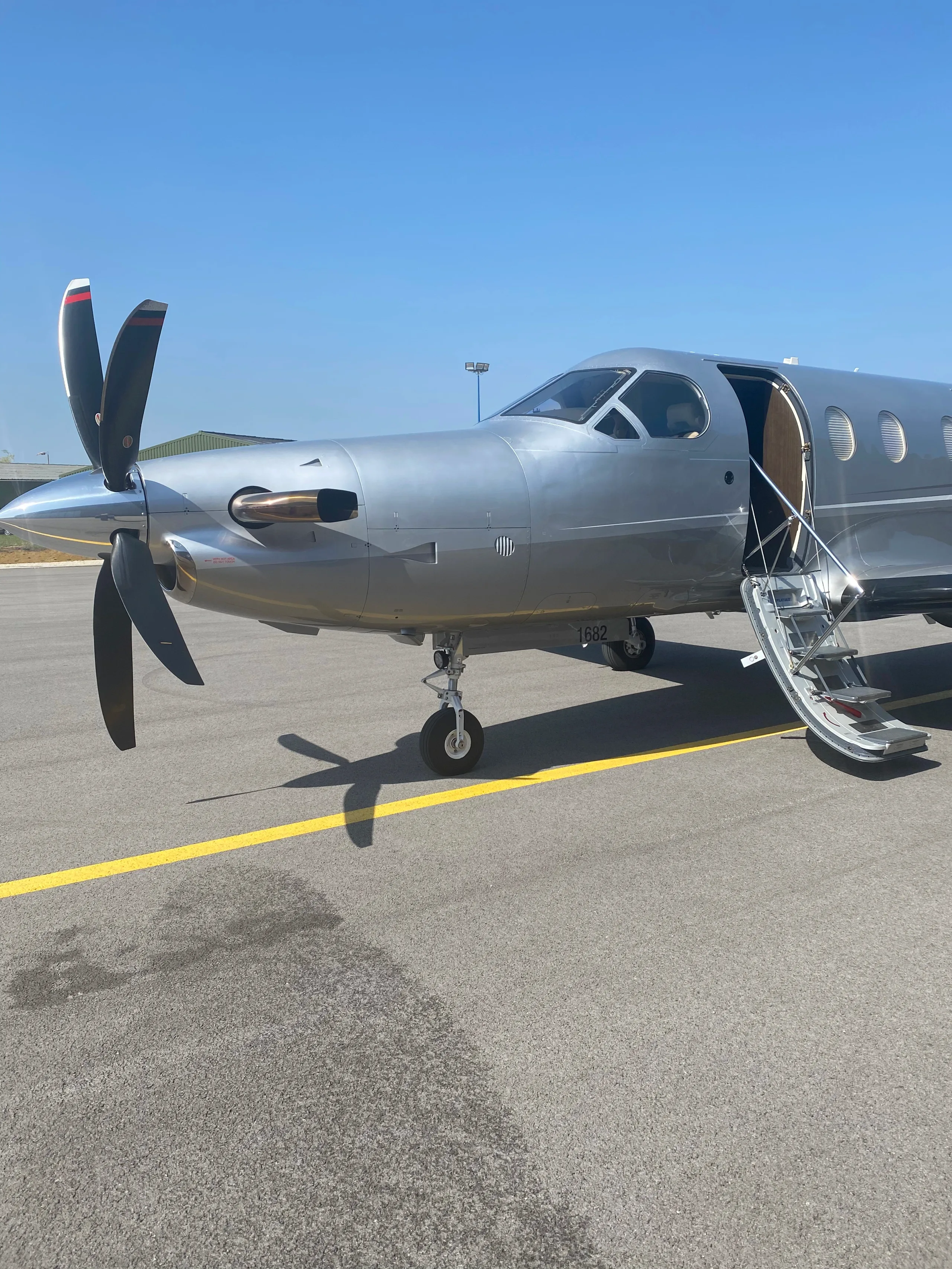 A close-up front-left view of a silver Pilatus PC-12 aircraft with its main door open and stairs extended, parked on an airfield under a clear blue sky.