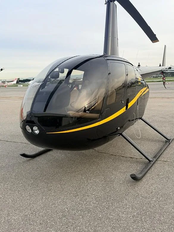 A black Robinson R44 helicopter with a yellow stripe on the side, parked on an asphalt tarmac.