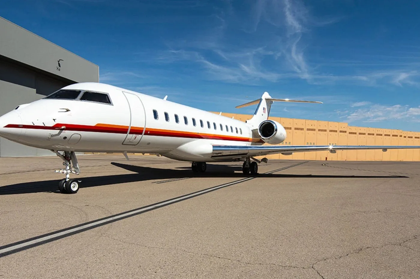 A white Bombardier Global business jet with red and orange stripes on the fuselage is parked on an airfield.