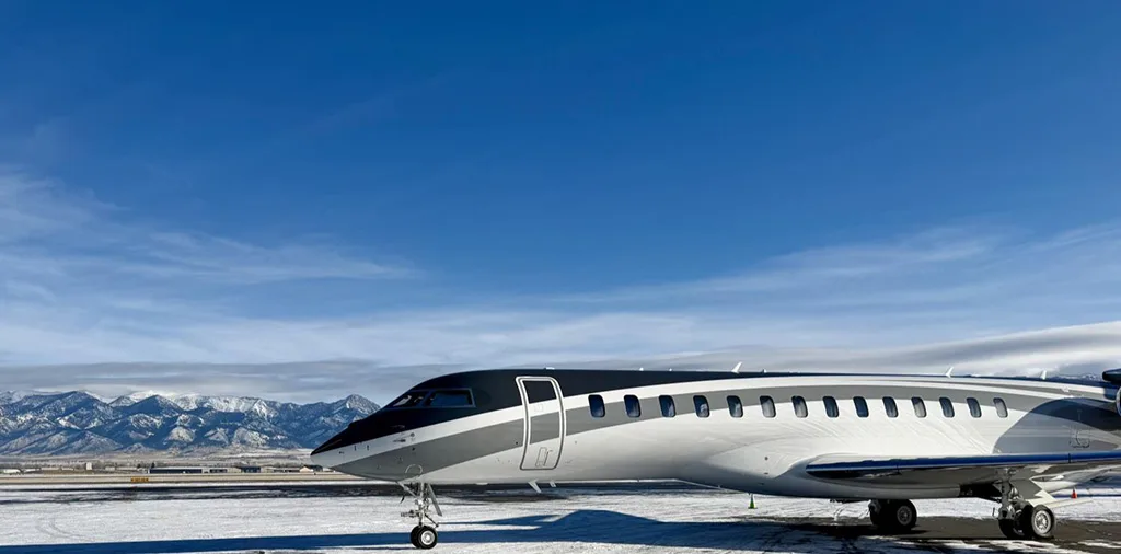 A grey and white private jet, likely a Global 7500, parked on a snowy tarmac with mountains in the background under a clear blue sky.