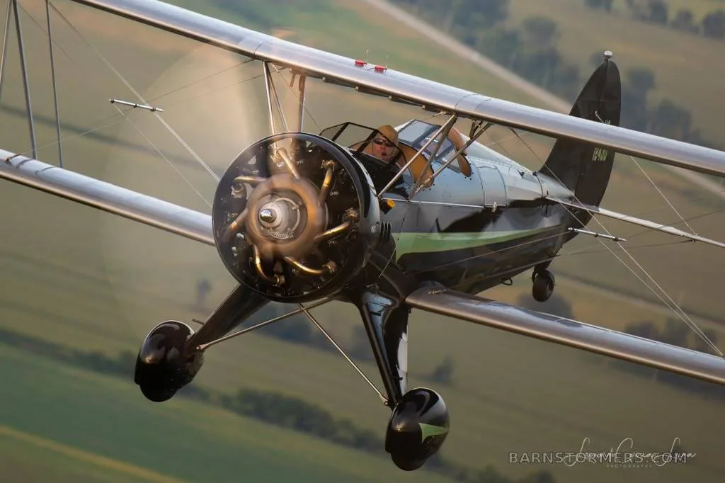 A black biplane with green accents and a radial engine is flying over green fields, seen from a low angle.