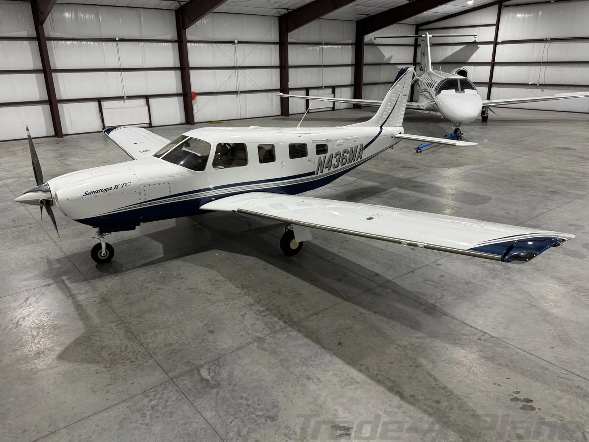 A white Piper Saratoga II TC N436MA with blue stripes is parked in a hangar with other aircraft.