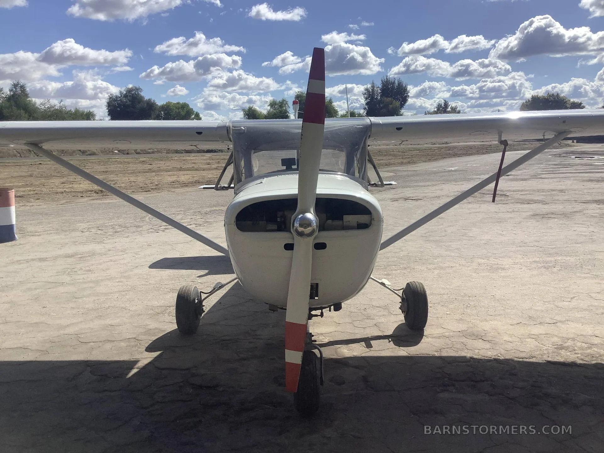 A front view of a white Cessna 150 with a red-tipped propeller, parked on a cracked asphalt surface under a blue sky with white clouds.