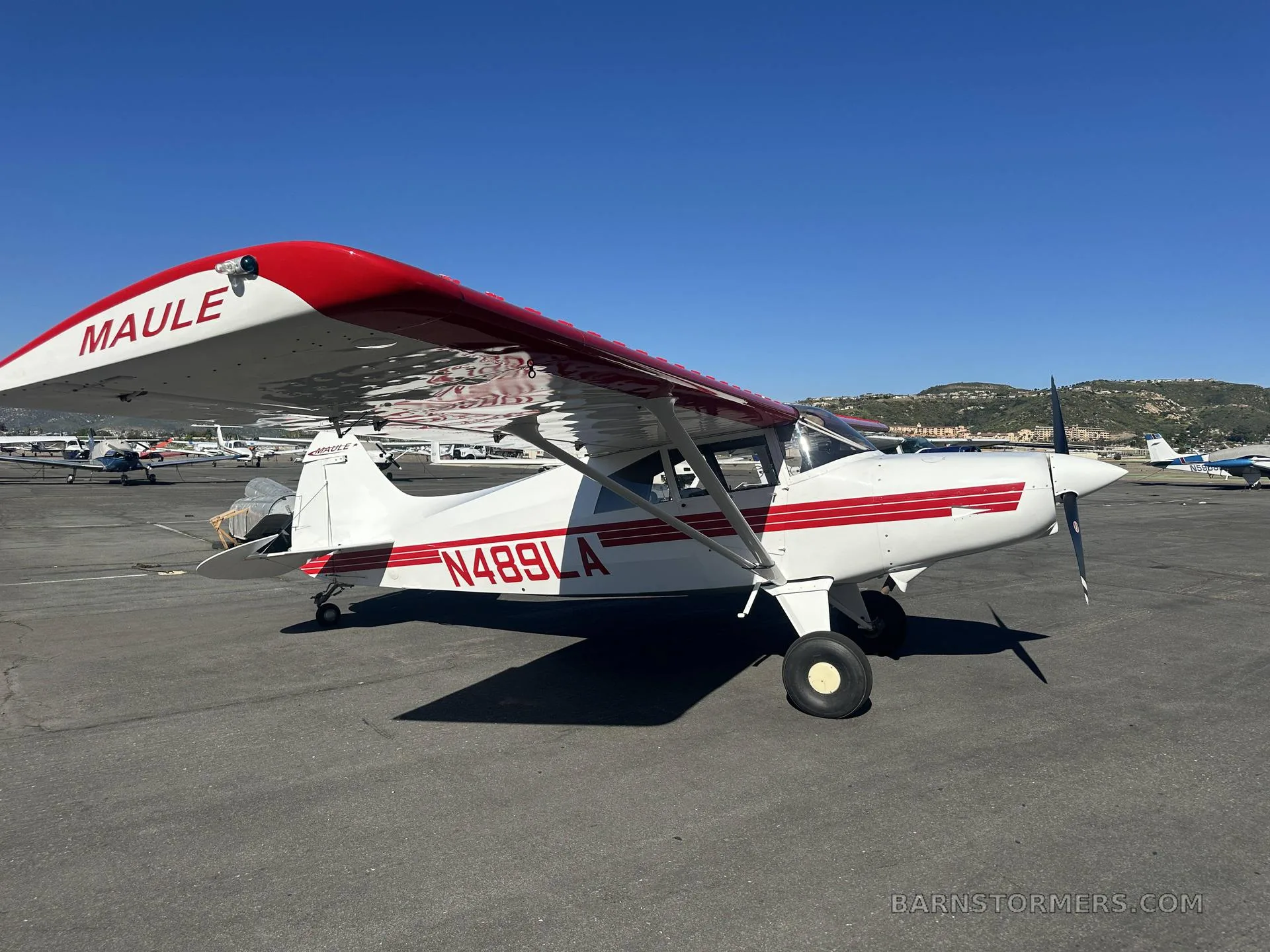 A white Maule M-7-235 aircraft with red stripes and registration N489LA is parked on an asphalt tarmac with hills in the background.