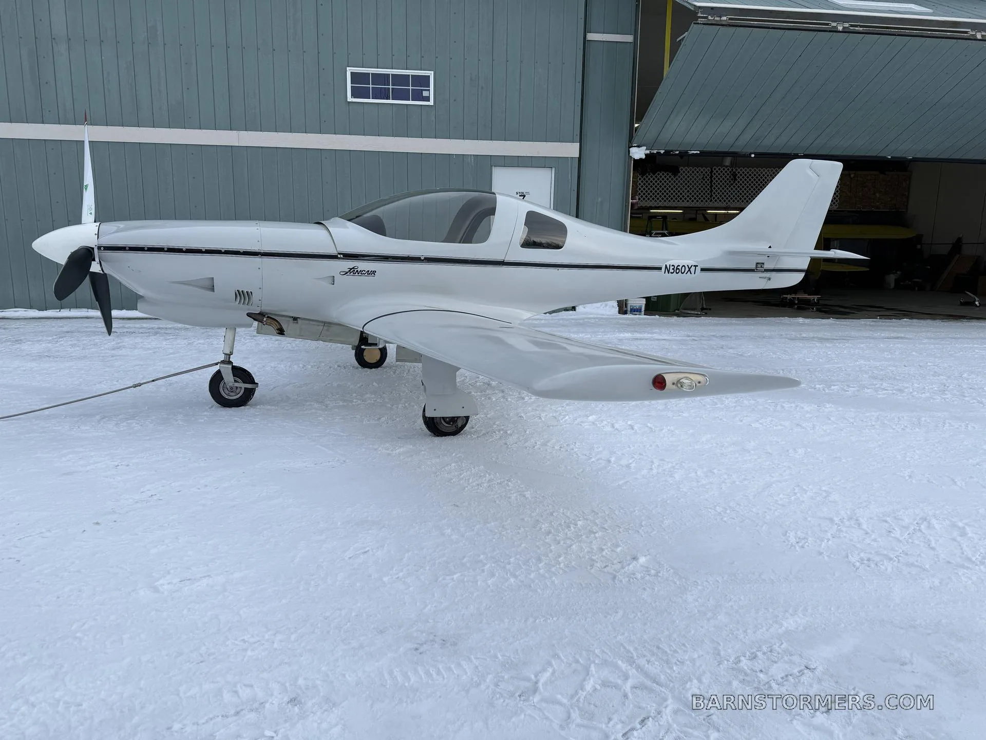 A white Lancair Legacy aircraft (N360XT) with a black stripe is parked on snowy ground in front of a blue hangar.