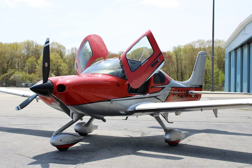 A red and silver Cirrus SR22 aircraft with registration N394JM parked on an tarmac with its gull-wing doors open.