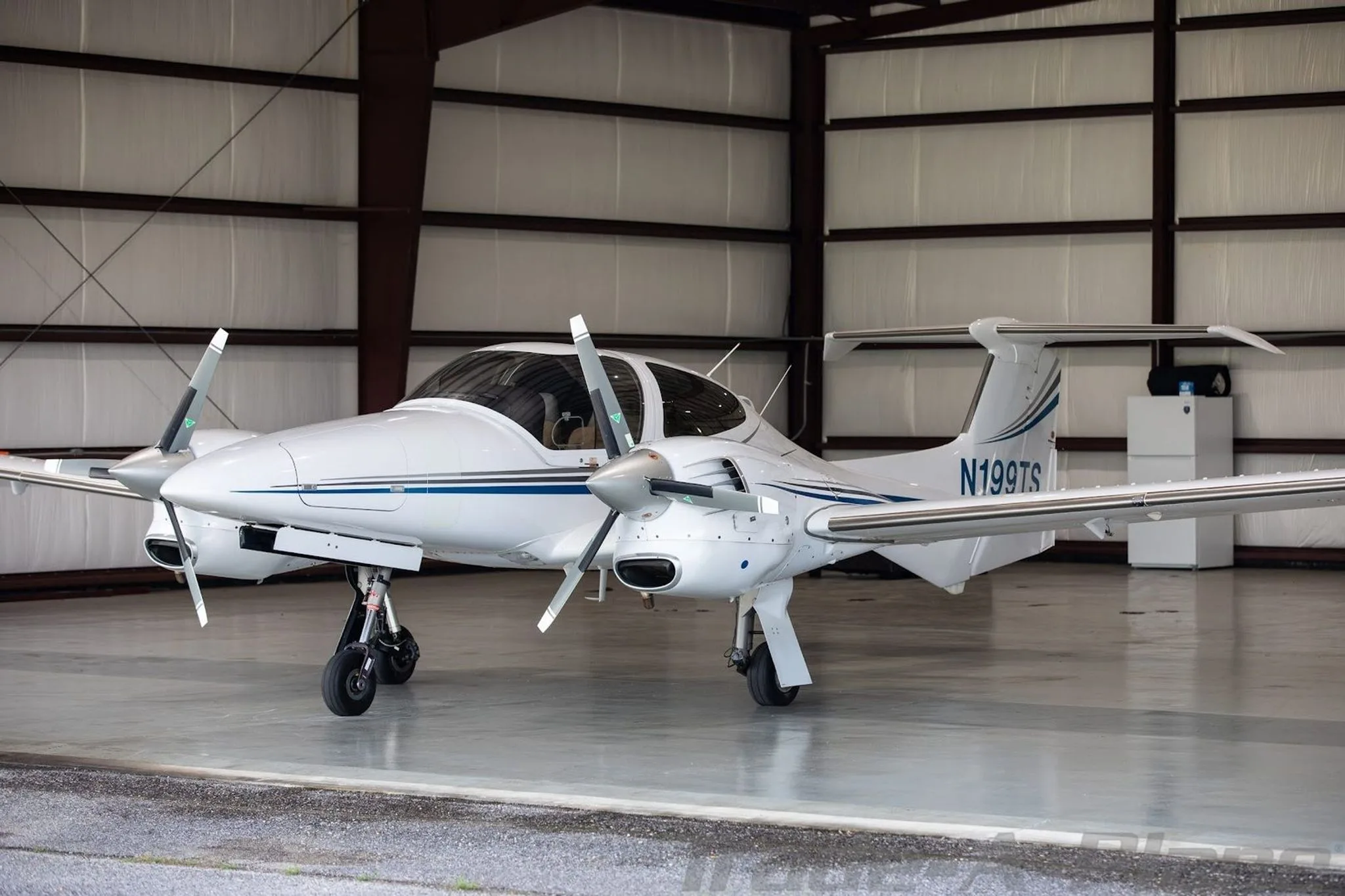 A white Diamond DA42-NG aircraft with blue and grey stripes is parked inside a hangar, viewed from the front-left.