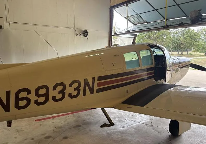 The side view of a white and brown Mooney M20 aircraft, registration N6933N, with its door open inside a hangar.