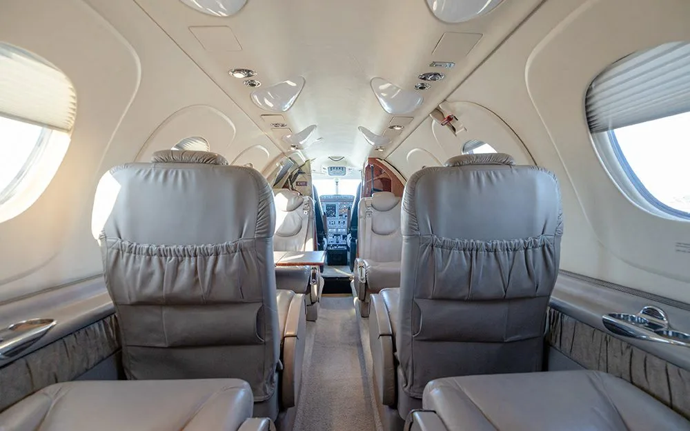 The beige interior cabin of a private jet, showing rows of seats and a view into the cockpit.