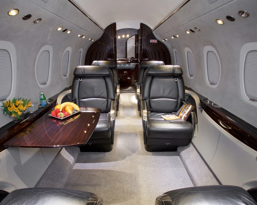 The spacious interior cabin of a private jet with black leather seats, a wooden table, and fresh fruit.