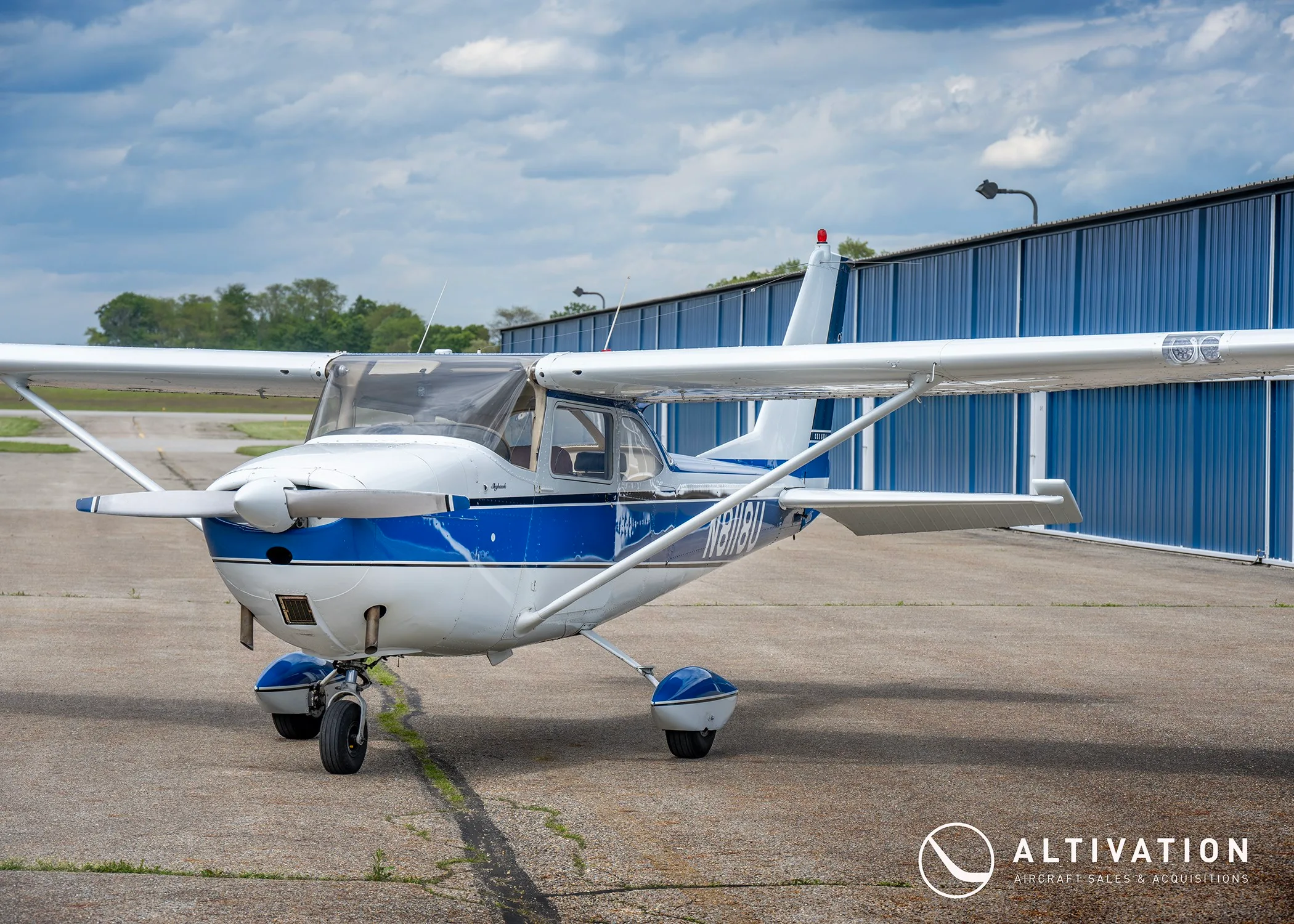 A front-quarter view of the white and blue Cessna 172 Skyhawk N8118U parked on an asphalt ramp next to a blue hangar.