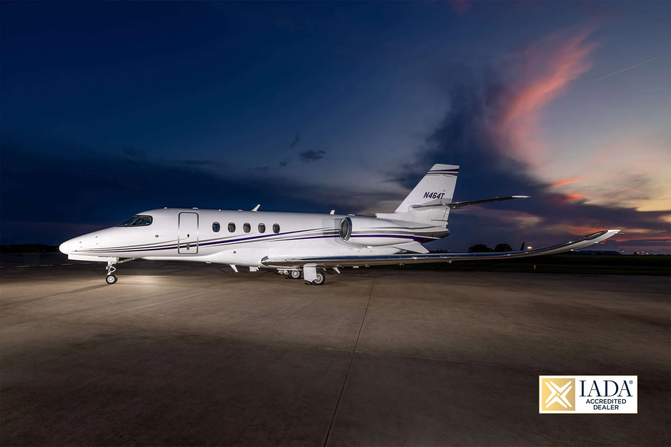 The N464T business jet is parked on an airfield at dusk, illuminated by its lights against a colorful sky.