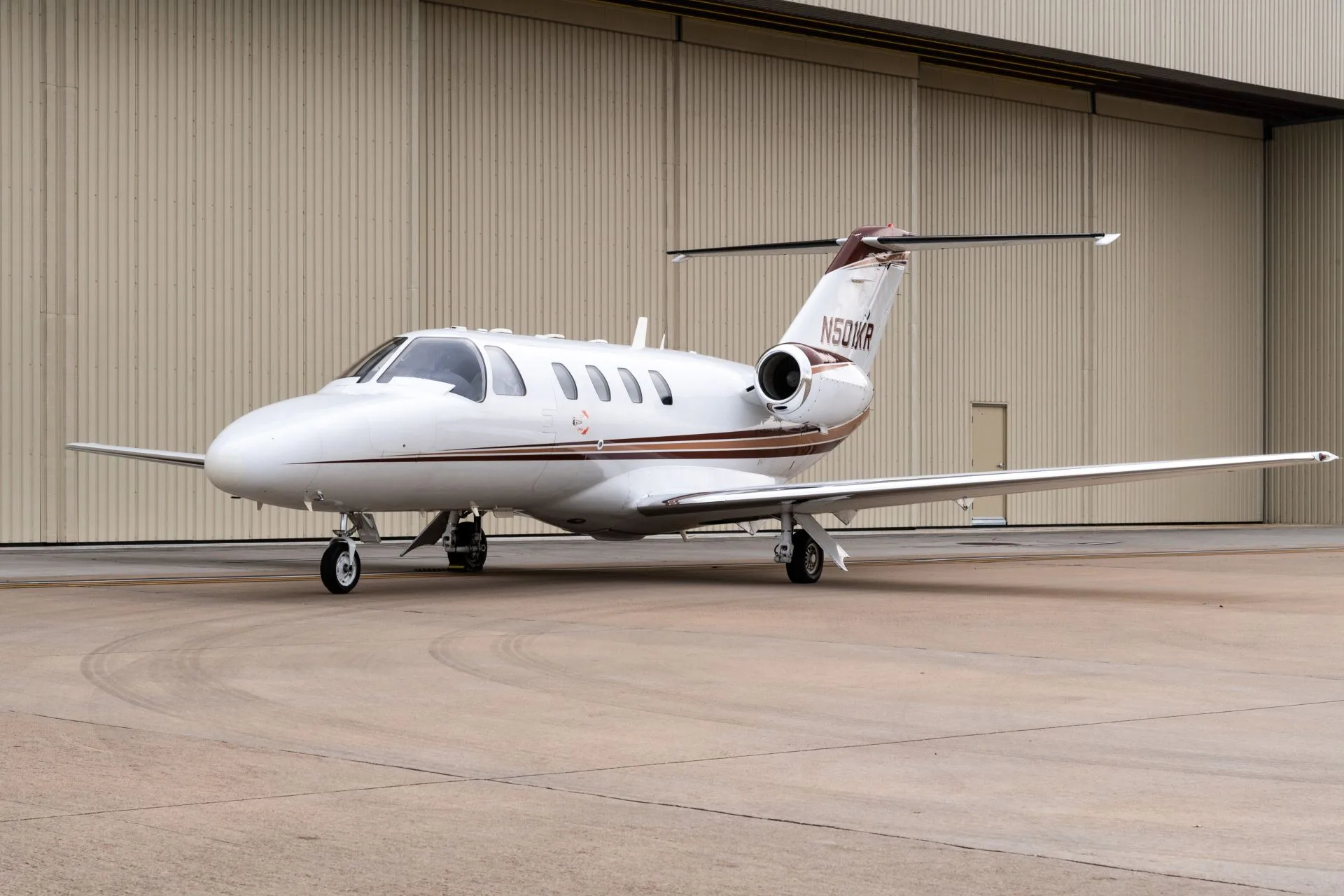 A white Cessna Citation jet with brown and gold stripes is parked on an asphalt apron in front of a beige hangar.