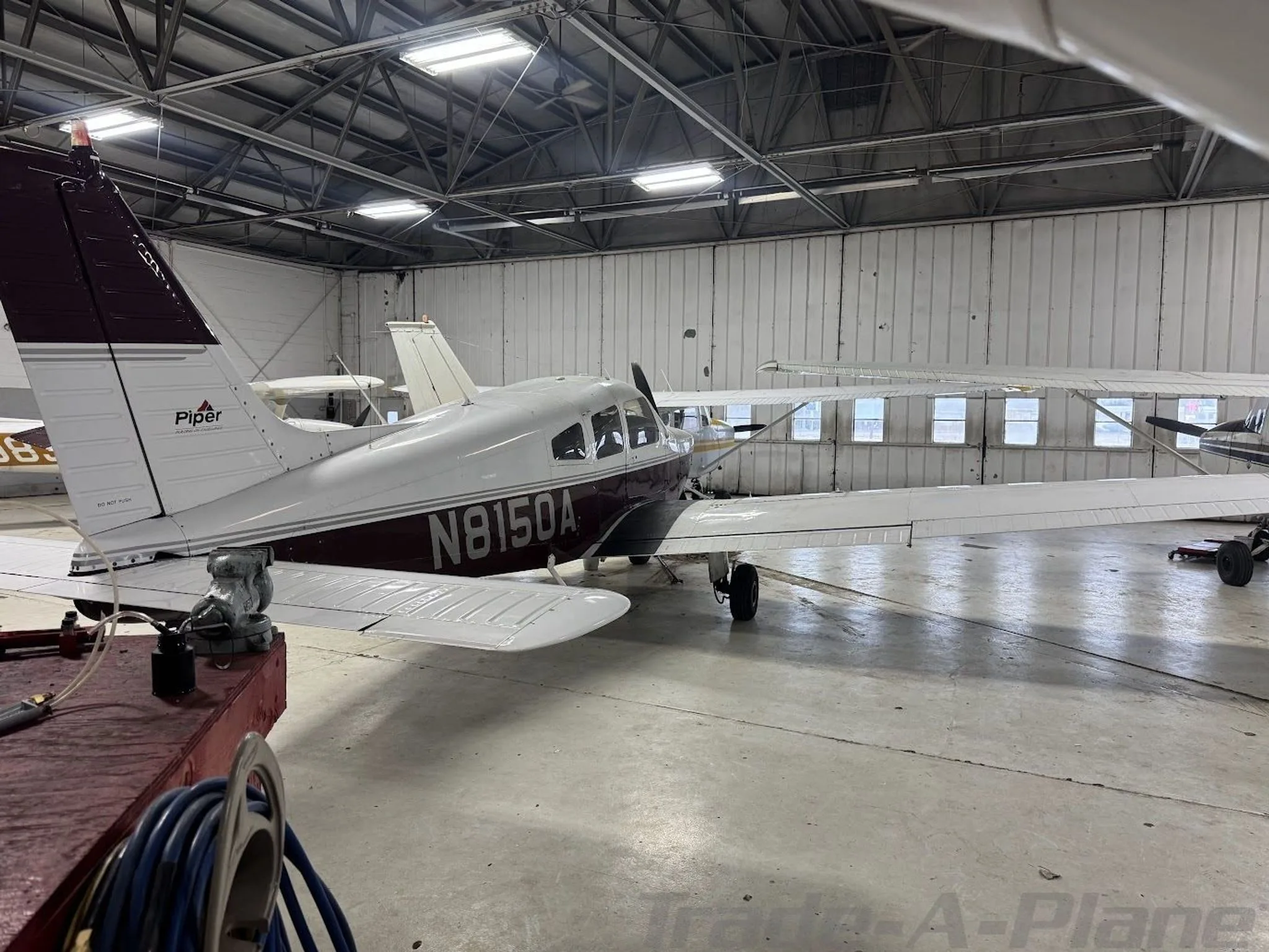 A white and maroon Piper Archer II (N8150A) parked in a hangar, with other aircraft in the background.