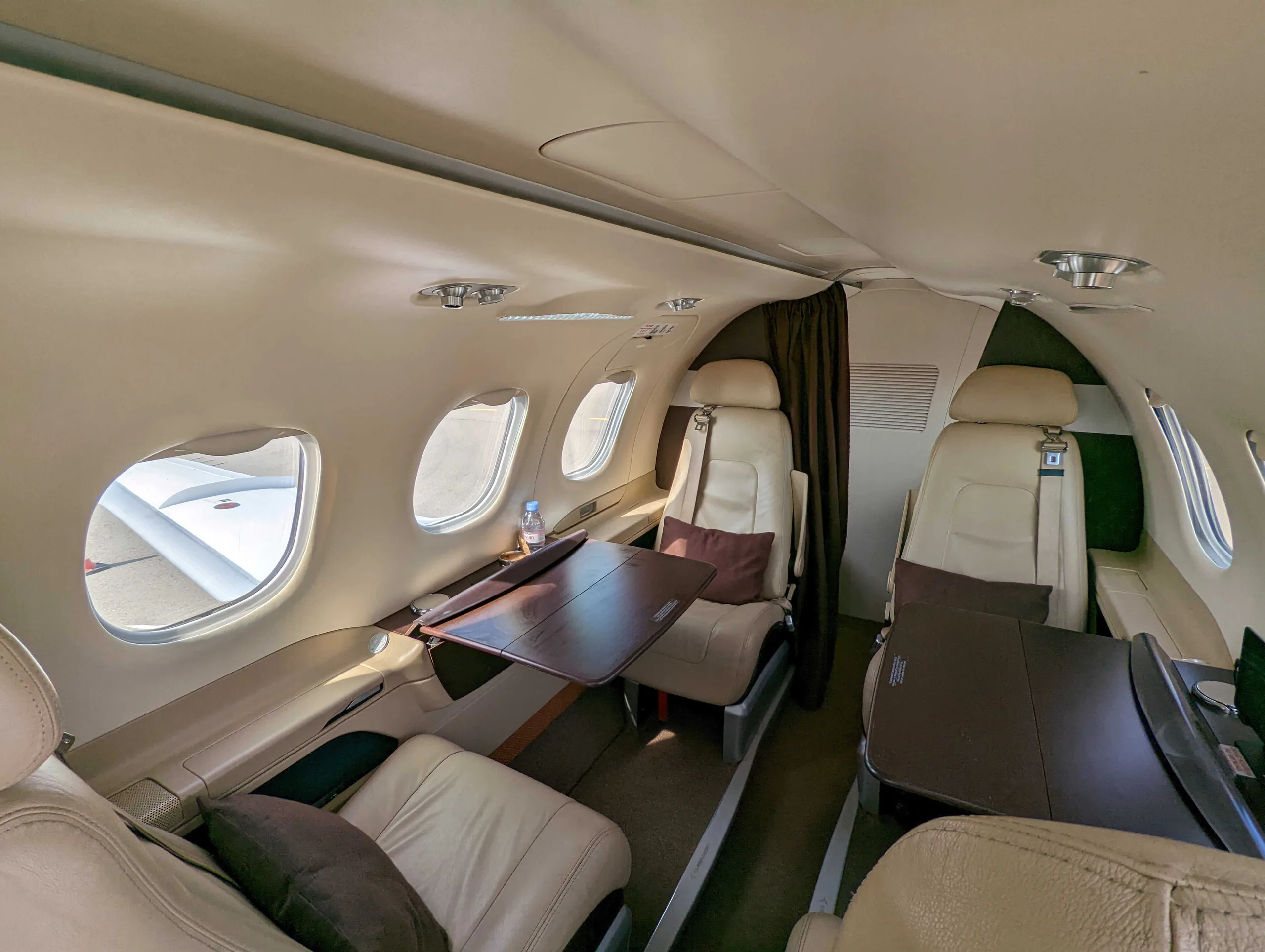 A view inside a private jet cabin shows beige leather seats, fold-out tables, and windows looking out onto an aircraft wing.