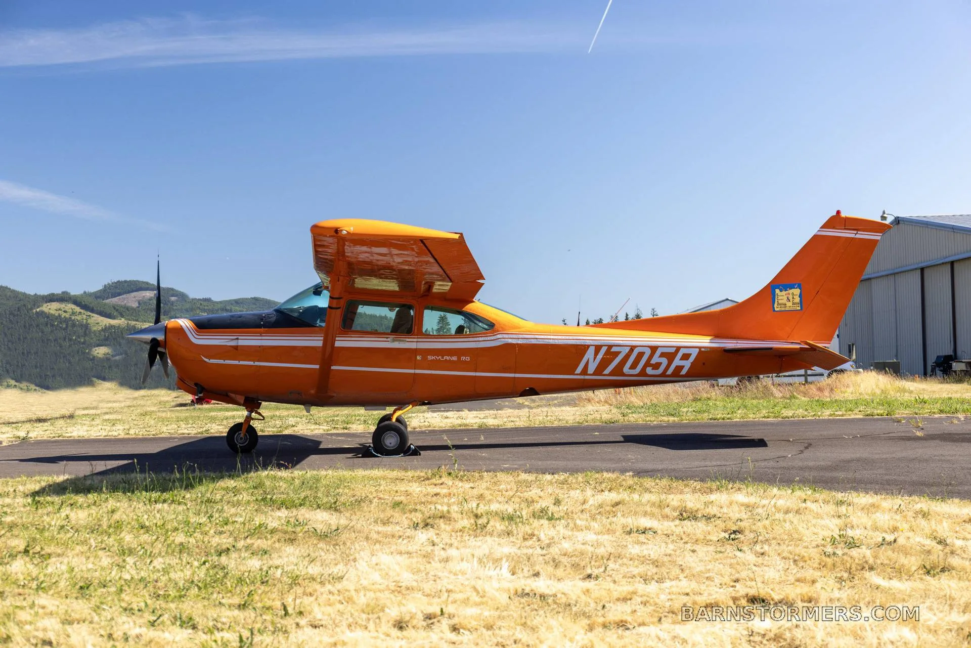 A bright orange Cessna 182 Skylane 50 with white stripes and black accents is parked on an asphalt strip next to a grassy field under a clear blue sky.