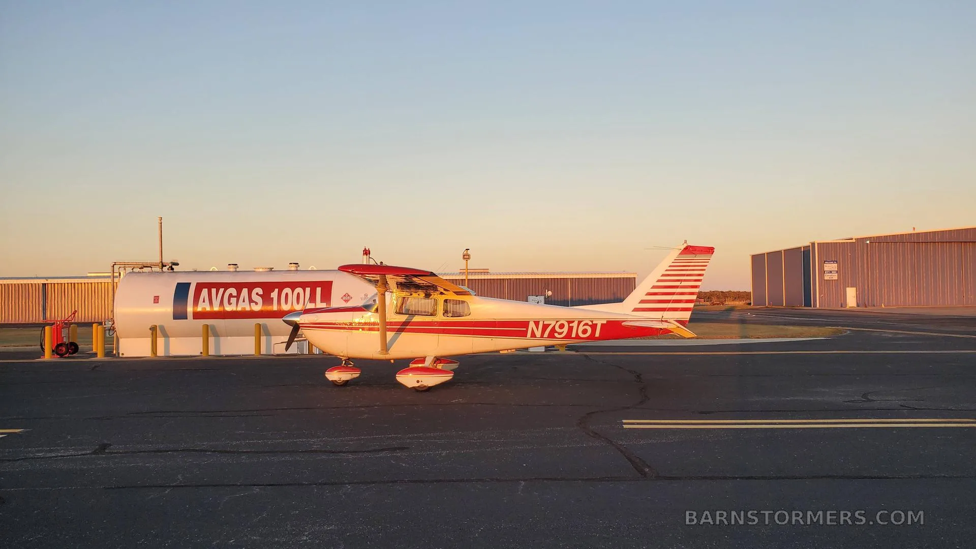 A white and red Cessna 172 with registration N7916T parked next to an AVGAS fuel tank at sunset.