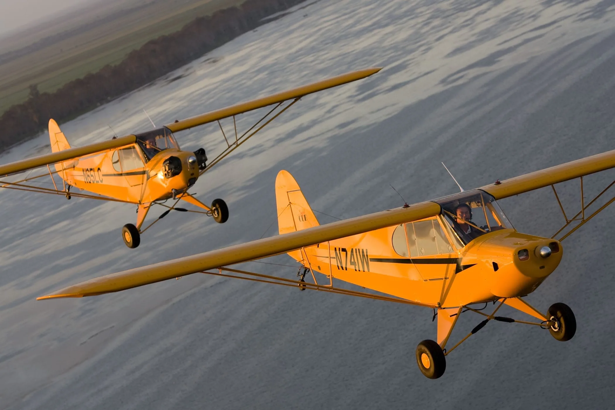 Two yellow Piper Cub-style aircraft (N741W and N65LC) fly in formation over water.