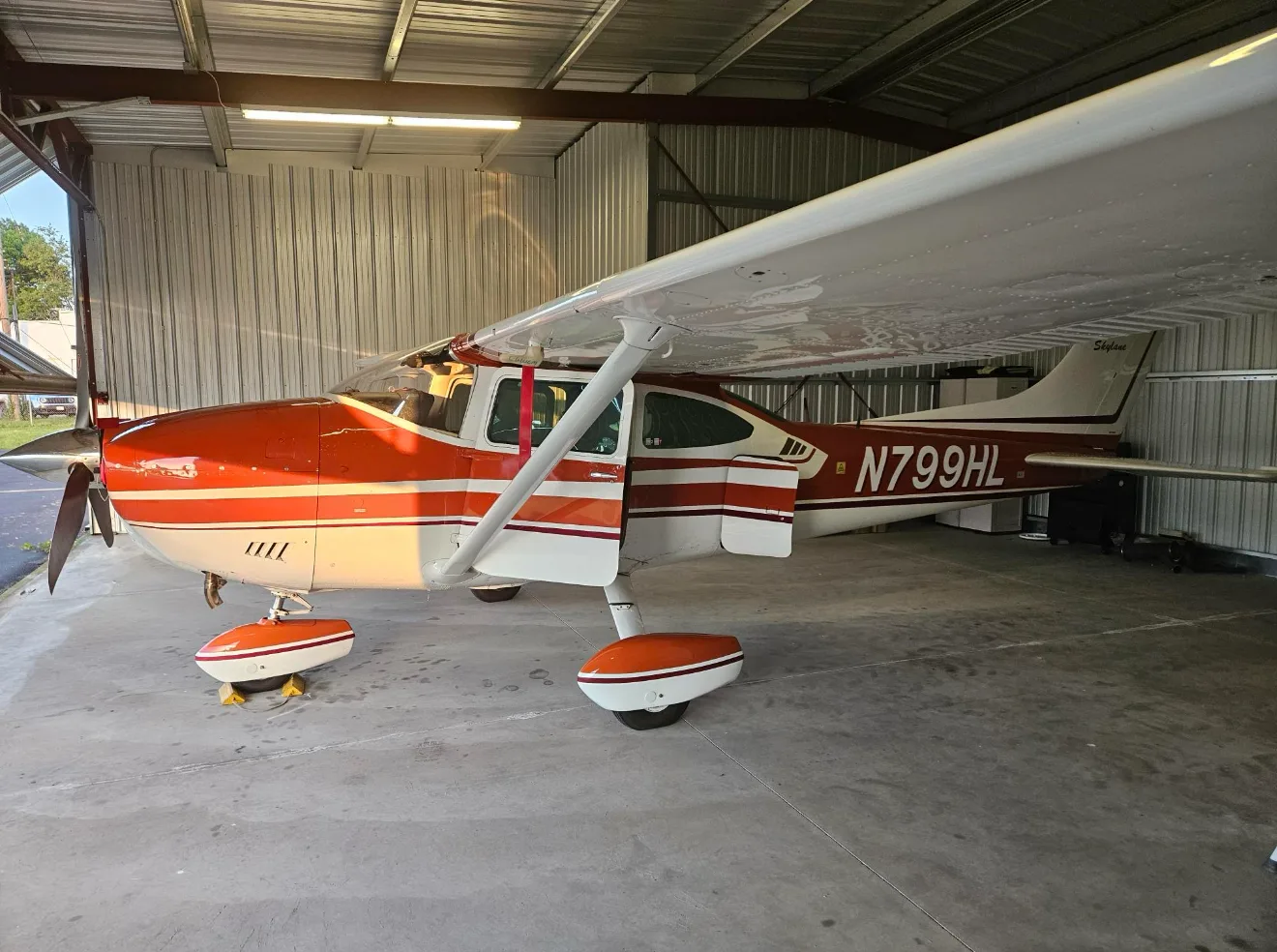 A red and white Cessna 182 Skylane with its door open is parked inside a hangar.
