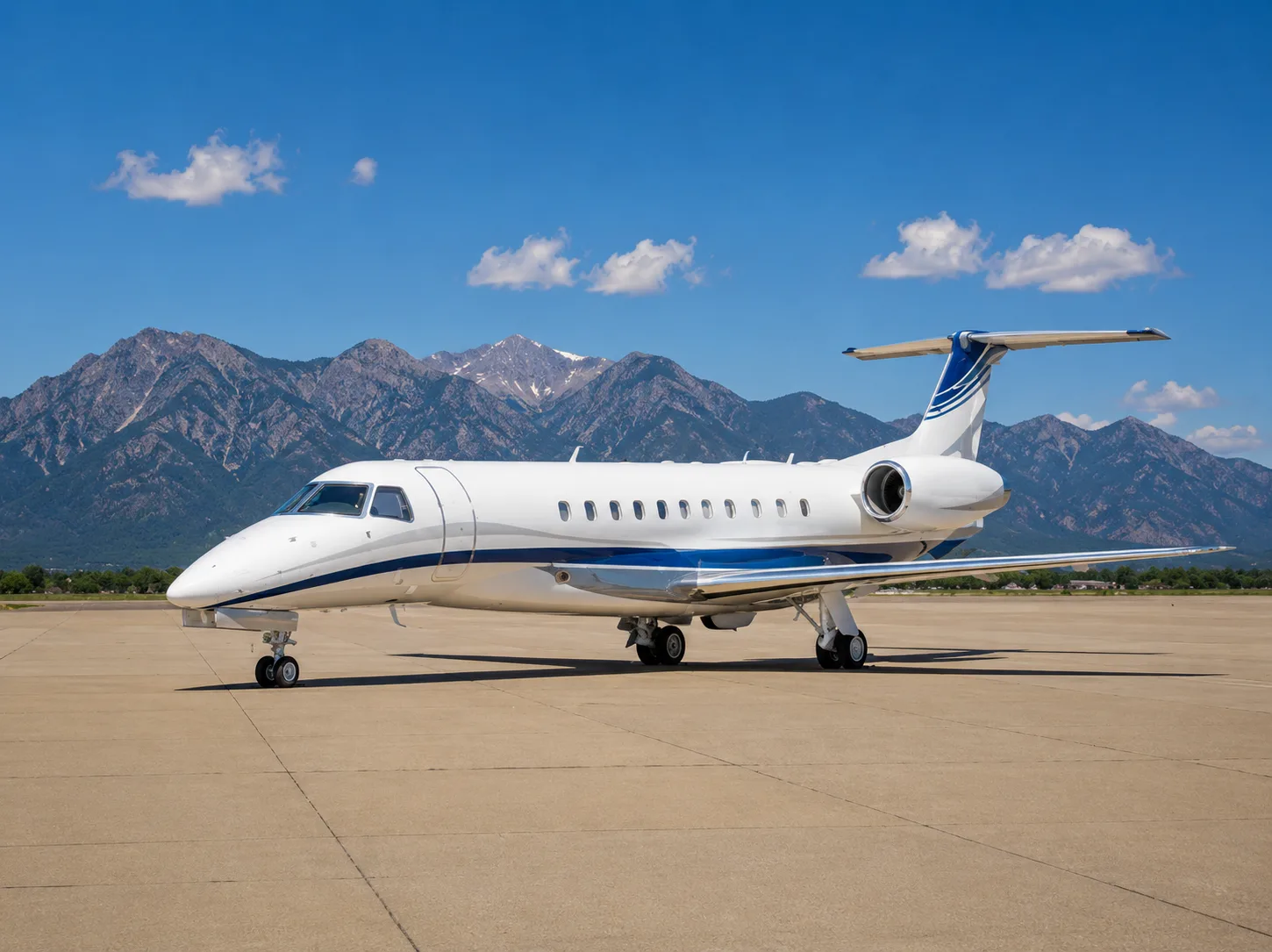 A white business jet with blue accent stripes is parked on an airfield with mountains in the background under a clear blue sky.