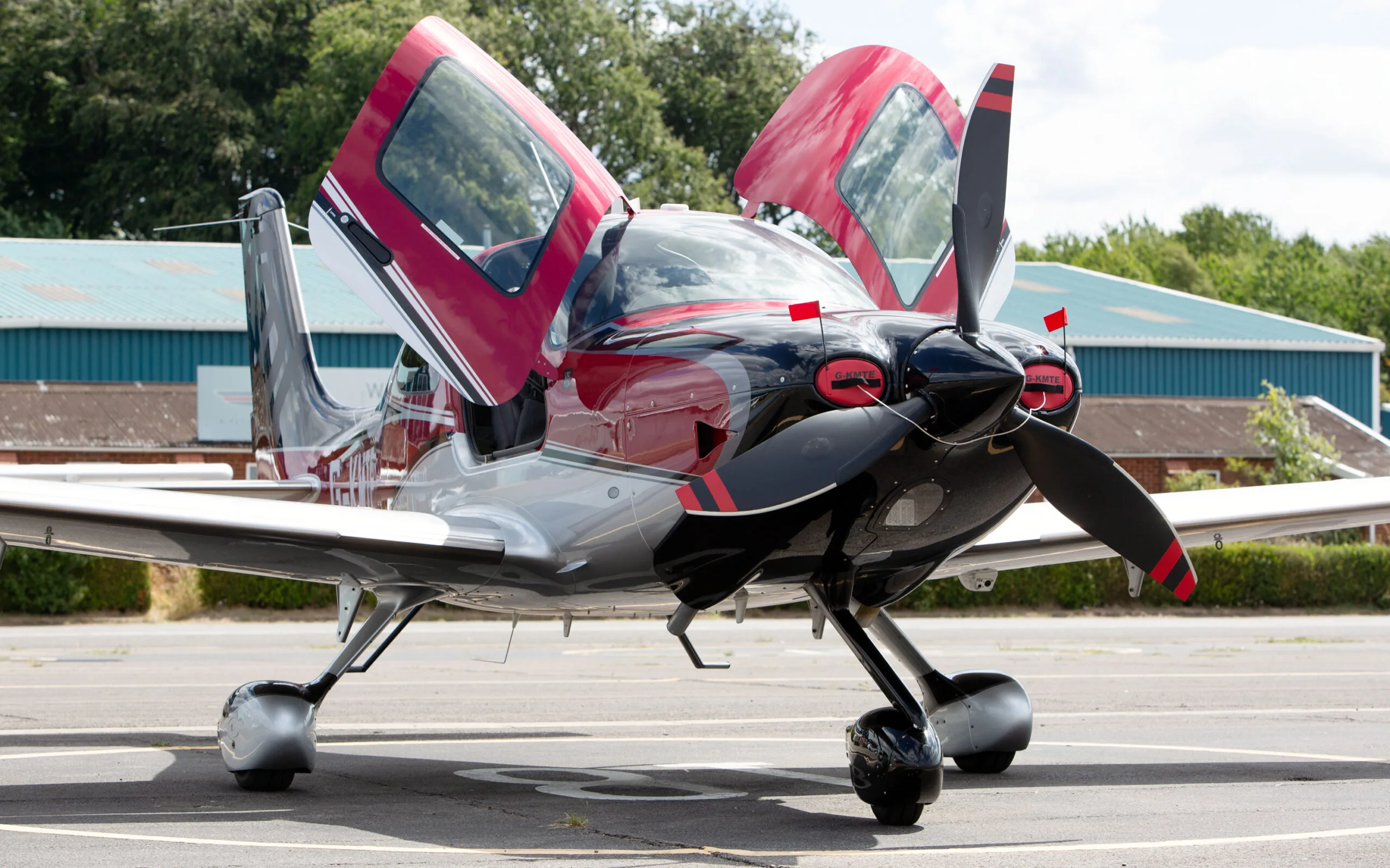 A front view of a red, black, and silver Cirrus SR22 aircraft with its gull-wing doors open, parked on an airfield.