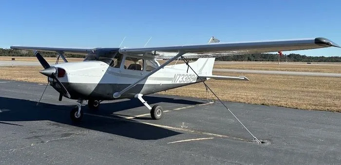 A white and gray Cessna 172 single-engine propeller aircraft is parked on an asphalt ramp at an airfield under a clear blue sky.