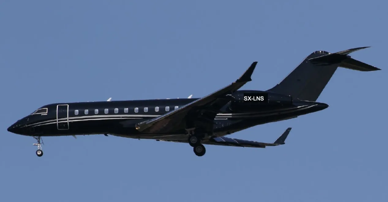 A black Bombardier Global Express XRS aircraft with registration SX-LNS in flight against a clear blue sky.
