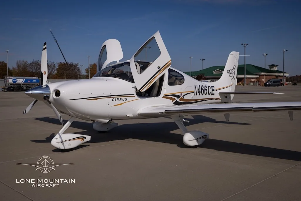 A white Cirrus SR22 aircraft with gold and black stripes, registration N466CE, parked on an airfield with its doors open.