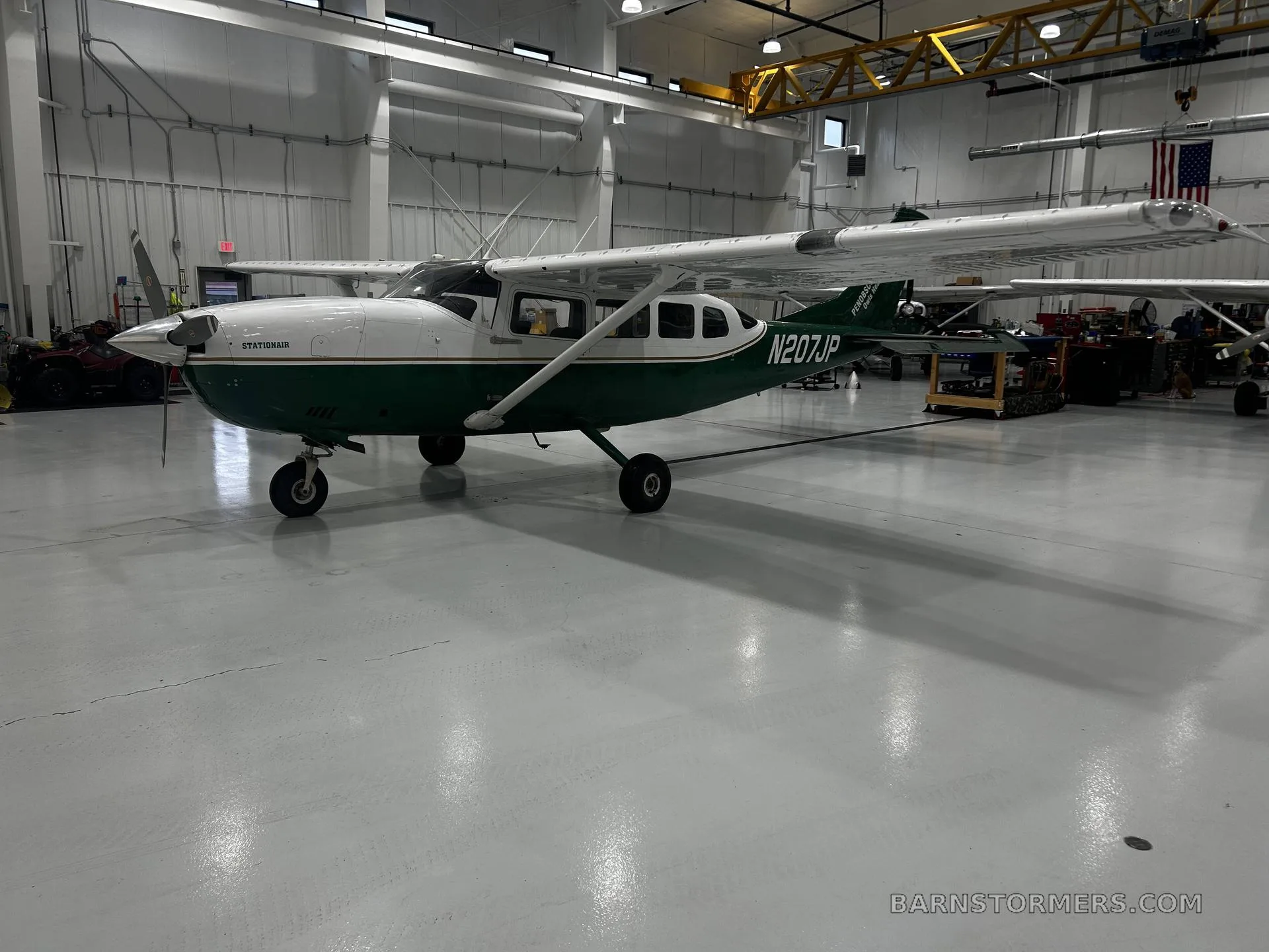 A white and green Cessna 207 Stationair, registration N207JP, is parked in a hangar, viewed from the front-right side.