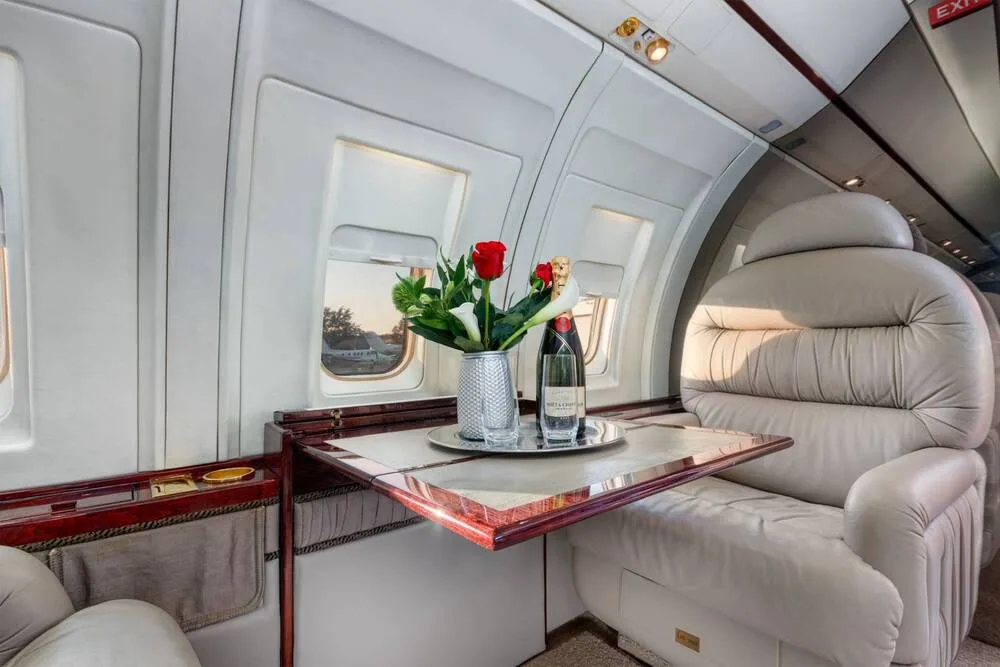 A luxurious aircraft interior showing a beige leather seat, a polished wooden table with flowers and champagne, and an exit sign above.