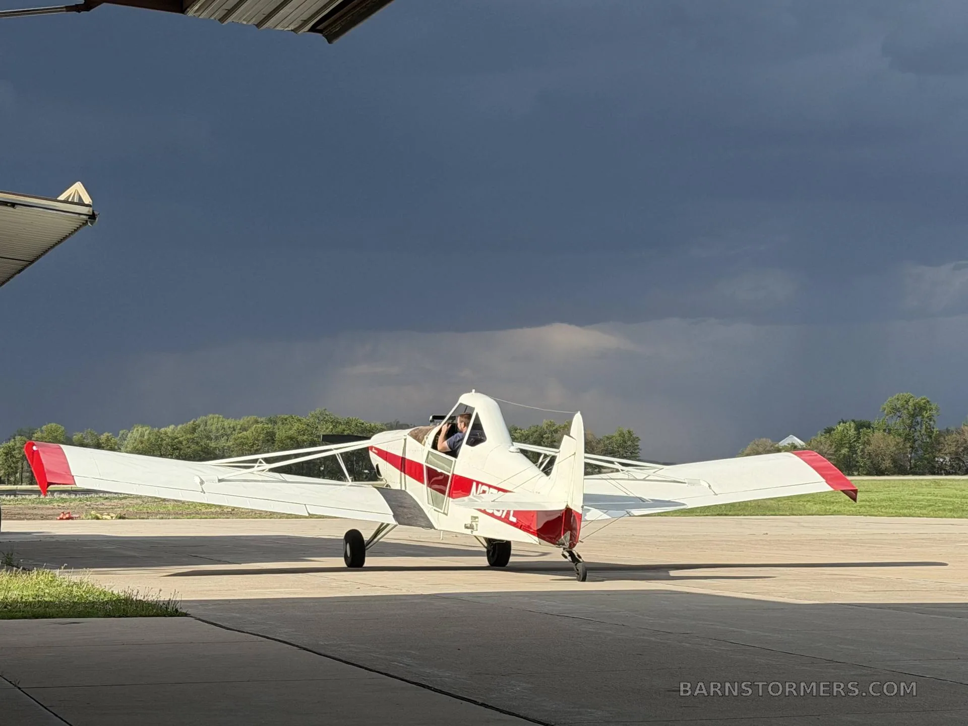 A white and red agricultural aircraft, seen from the rear-left, with a pilot in the cockpit, parked on an airfield under a dark, cloudy sky.