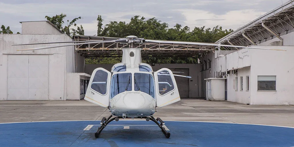 A white helicopter with gold stripes is parked on a blue helipad with all its doors open, viewed from the front.
