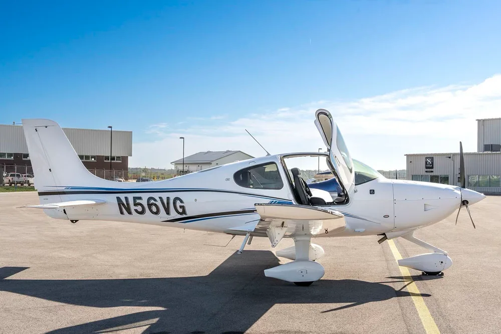 A side view of the white Cirrus SR22 aircraft with blue and black stripes, tail number N56VG, parked on an airfield with the pilot's door open.