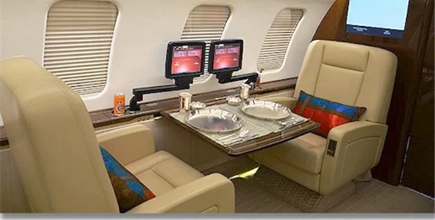 A luxurious aircraft interior with two beige leather seats facing each other, a dining table set for two, and dual entertainment screens mounted on an arm.