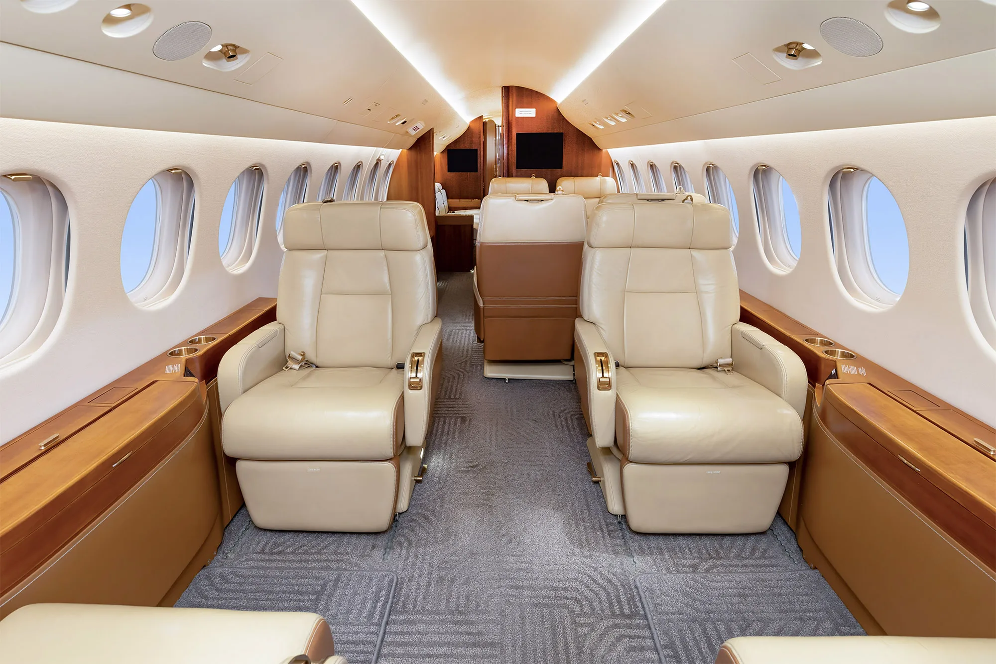 The luxurious cabin interior of a business jet, featuring two rows of beige leather seats facing each other, with wood paneling and large windows.