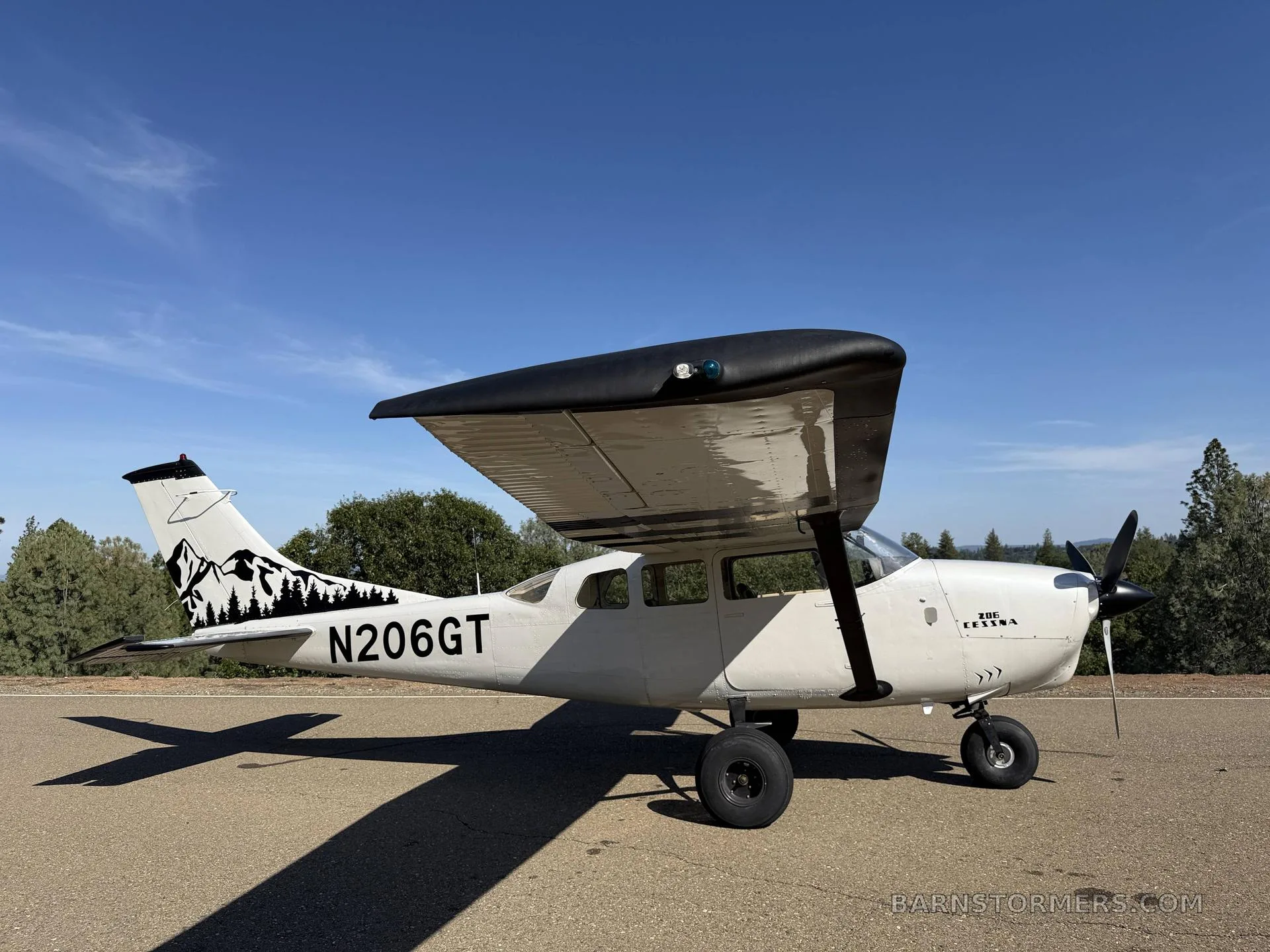 A side view of a white Cessna 206GT with black wings and mountain tail art, parked on a paved surface.