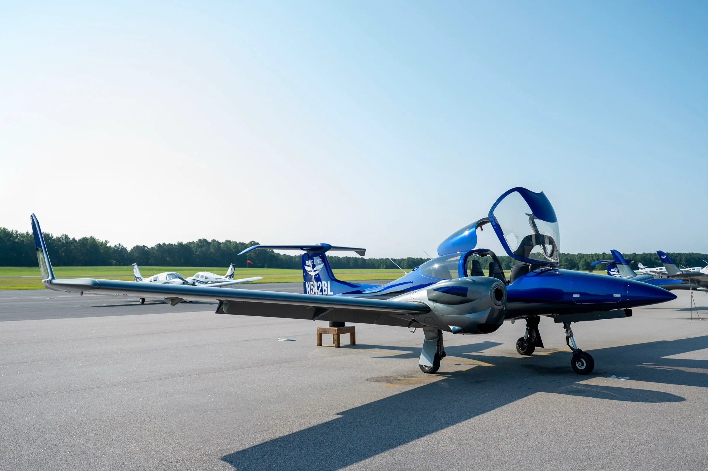 A blue and grey Diamond DA42 aircraft with doors open, parked on an airfield with other aircraft in the background.
