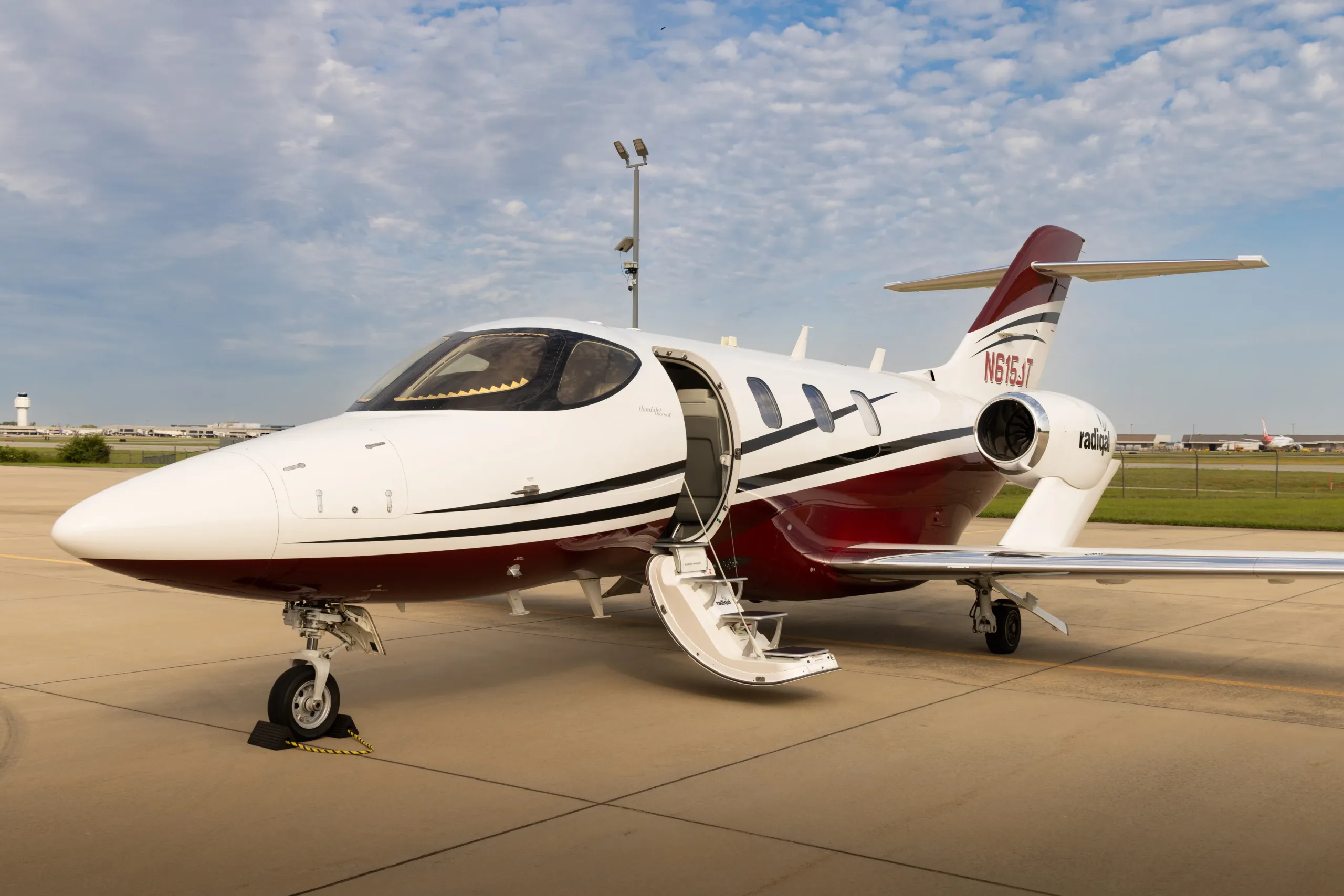 A white and maroon HondaJet Elite II (N615JT) with its door open, parked on a tarmac under a cloudy sky.