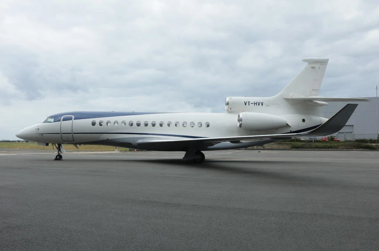 A white and blue Dassault Falcon 900 business jet with registration VT-HVV parked on an airfield under a cloudy sky.